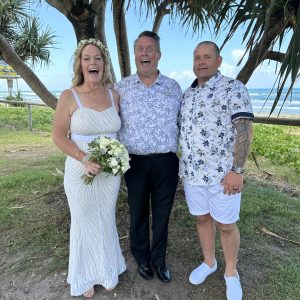 Mark Reynolds Celebrant posing with wedding couple laughing with beach in the background.
